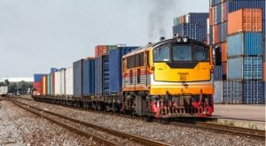 A photo of a train transporting shipping containers with additional stacks of shipping containers in the background.