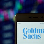 The Goldman Sachs logo is displayed on a smartphone in front of a multi-color background.