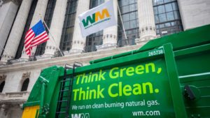 Image of green Waste Management (WM) branded truck in the foreground and building with Waste Management flag in the background.