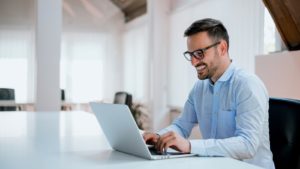 a man in a collared shirt smiles while typing on his laptop