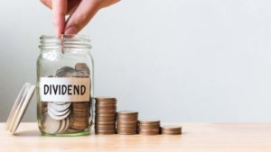 Hand putting coin in jar word dividend with money stack.