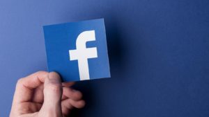 Facebook (FB) logo held by hand backdropped by company-blue background
