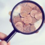 A hand holding a magnifying glass over pennies. under-$1 stocks to buy