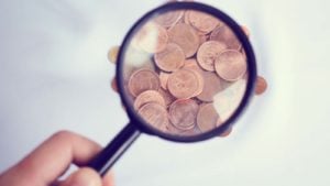 A hand holding a magnifying glass over pennies. under-$1 stocks to buy