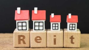 tiny house figures atop letter blocks spelling out REIT, representing reits to buy. stock predictions. best REITs