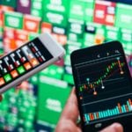 Low-beta stocks: traders use smartphones to trade stocks in front of a wall of green and red tickers