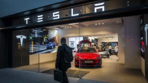 A person walks past the storefront of a Tesla store with several vehicles visible behind a glass door