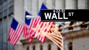 Street sign for Wall Street pictured in front of several US flags depicting US stocks