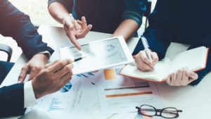 Three people sit around a table holding financial charts and a tablet device.