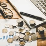 A laptop, pencil, pair of eyeglasses, and many coins rest on a wooden table. financial stocks to buy