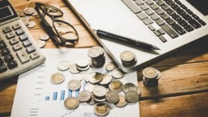 A laptop, a pencil, glasses and a lot of coins lie on a wooden table.