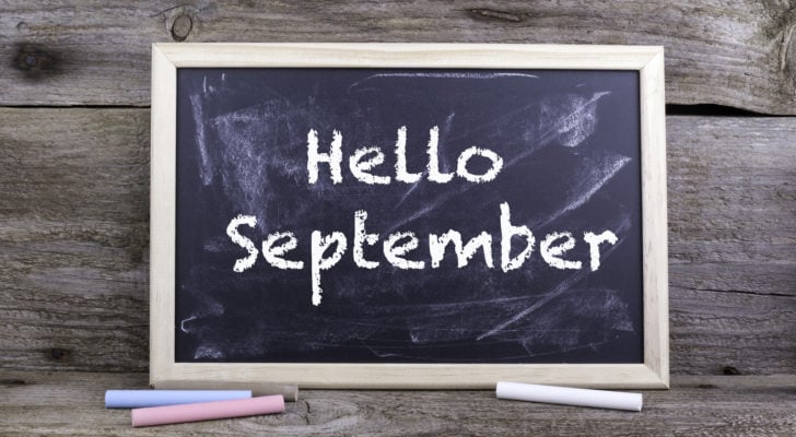 An image of a rectange chalkboard leaning on grey wood planks with the text "Hello September" in a white chalk style font with a pink, blue, brown, and white chalk stick laying in front.