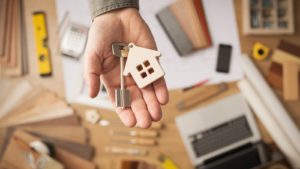 Image of a man holding a keychain with a key and a house on the keyring above an office desk in the background