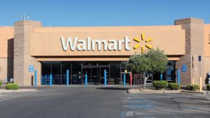 An image of a large tan Walmart store front on a bright clear day with empty handicap parking spaces, trees, bushes, and blue poles in front and a large white "Walmart" sign and logo with six yellow lines forming a circle above the entrance.