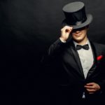 An image of a man wearing a black and white tuxedo using his right hand to tip his black top hat in front of a black background.