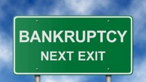 A green road sign reads "Bankruptcy Next Exit" in front of a cloudy blue sky.