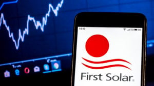 A smartphone showing a red circle, two red curvy lines, and the text "First Solar" on a white screen, with a computer screen showing a Windows taskbar and a blue and white stock chart in the background.