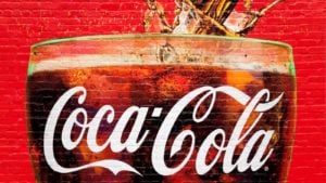 An image of a large brick wall painting of the top of a glass with a white "Coca-Cola" logo on it filled with soda and ice with soda splasing from the top of it on a red background.