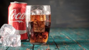 An image of three square ice cubes stacked up, a red Coco-Cola can, and a glass with ice and soda (from left to right) on a teal painted wood table.