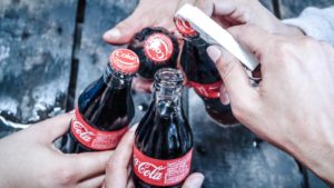 Close-up photo of hands holding glass Coca Cola (KO) bottles, clinking them together. One hand has a bottle opener and is opening a bottle.