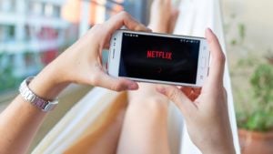 Picture of a person laying on a couch holding a mobile phone that features the Netflix (NFLX) logo on the screen