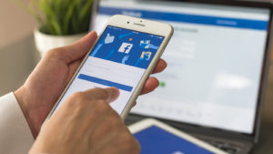 image of person using a Facebook (FB) app on their mobile phone with Facebook also open on an online browser in the background