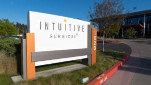 晴れた日に、白、グレー、オレンジの建物の前の芝生の上にある、「Intuitive Surgical」のロゴと番号「1020」が記載された白、グレー、オレンジの看板の画像。