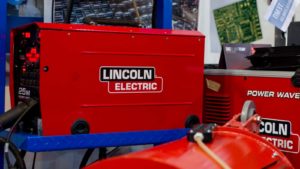 An image from a mechanical shop with a red Lincoln Electric wire feeder and power wave machine, with posters on a white wall in the background.