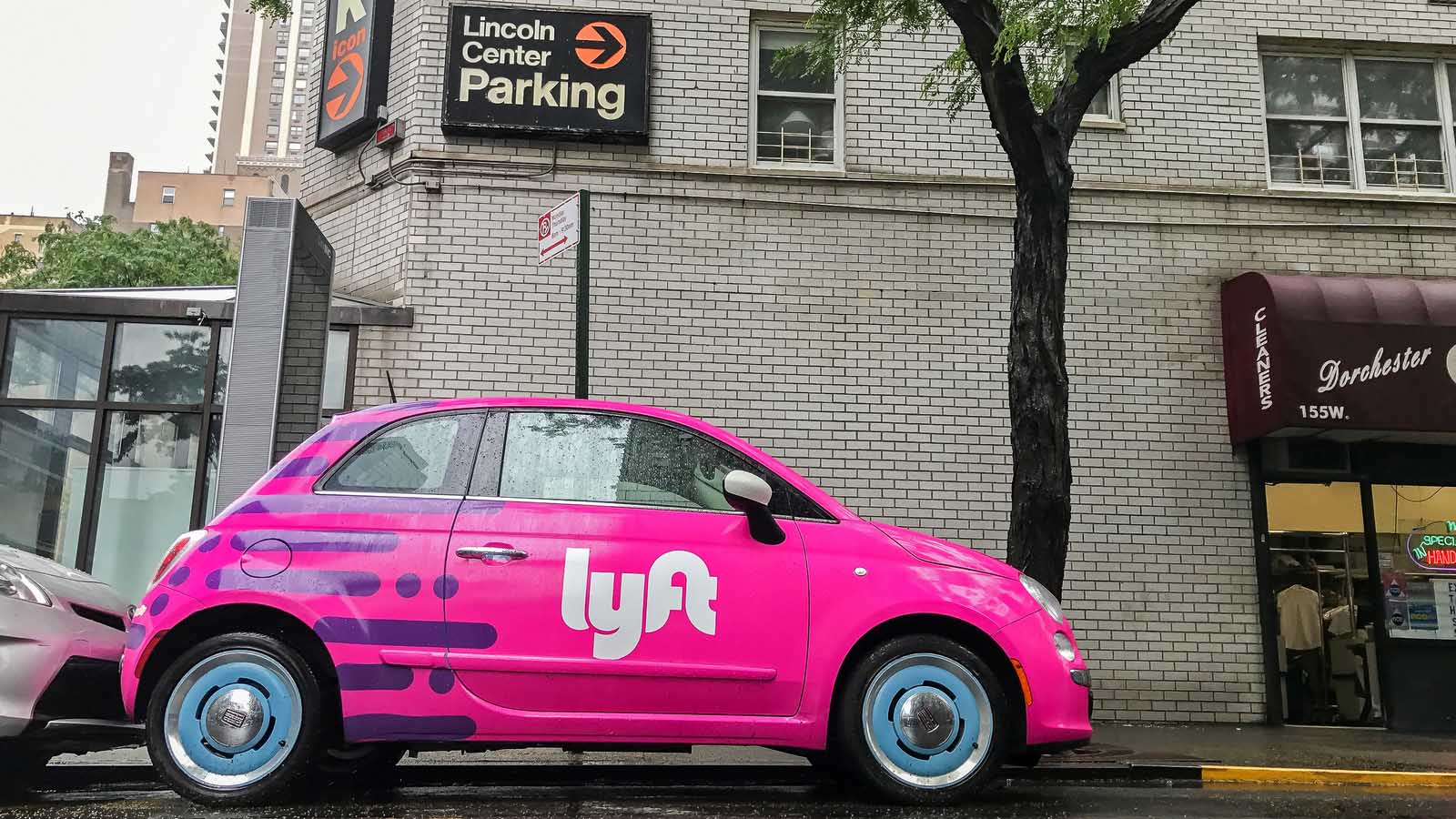 The Lyft (LYFT Stock) logo on the side of a pink car parked on a street.