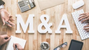 The letters "M&A" on top of a wooden table surrounded by hands, a keyboard, notepad and calculator.