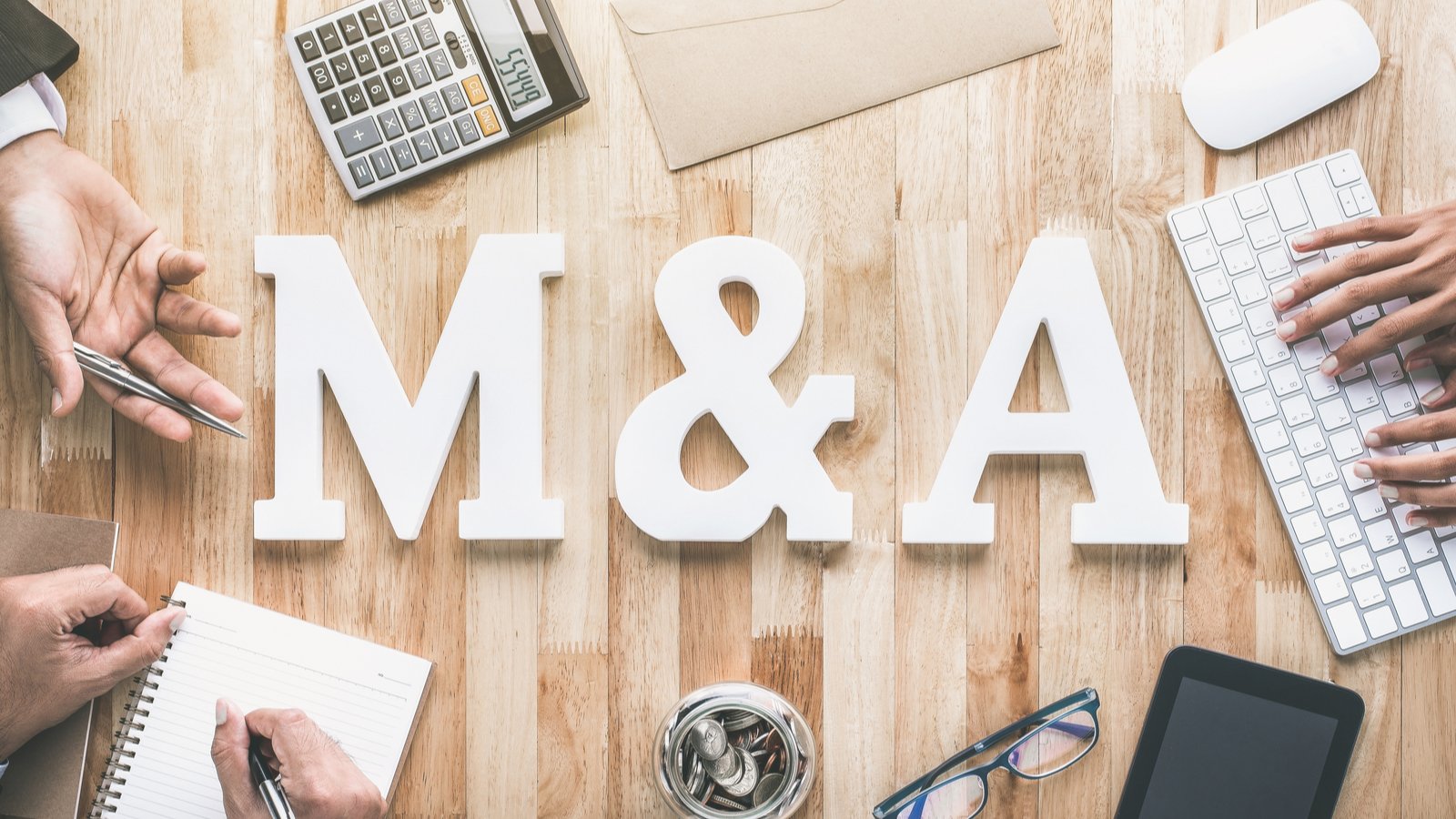 The letters "M&A" on a wooden table, surrounded by a calculator and other business items. TGA Stock