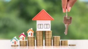 Toy houses rest atop stacks of coins while a hand dangles a set of keys in the air.