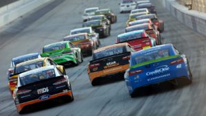 An image from behind around twenty race cars of various colors in two uneven rows racing on a race track.