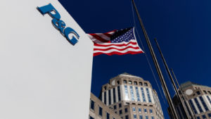 An image of a tan building corner with a blue "P&G" logo, an American flag on a post, and two larger building tops in the background under a dark blue sky.