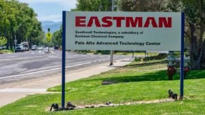 An image of a lawn in front of the street on a sunny day, with a white sign that has a red "Eastman" logo and black text beneath it, and fire hydrants, cars, and trees in the background.