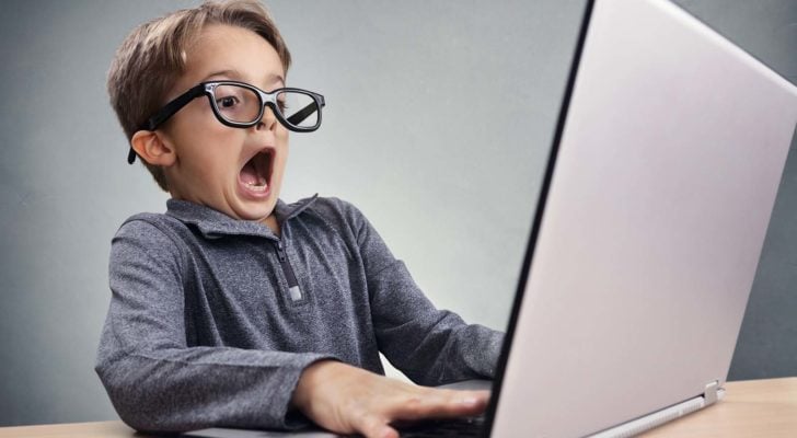 An image of a young boy with a grey sweater and black glasses sitting with a laptop at a table, looking very shocked.