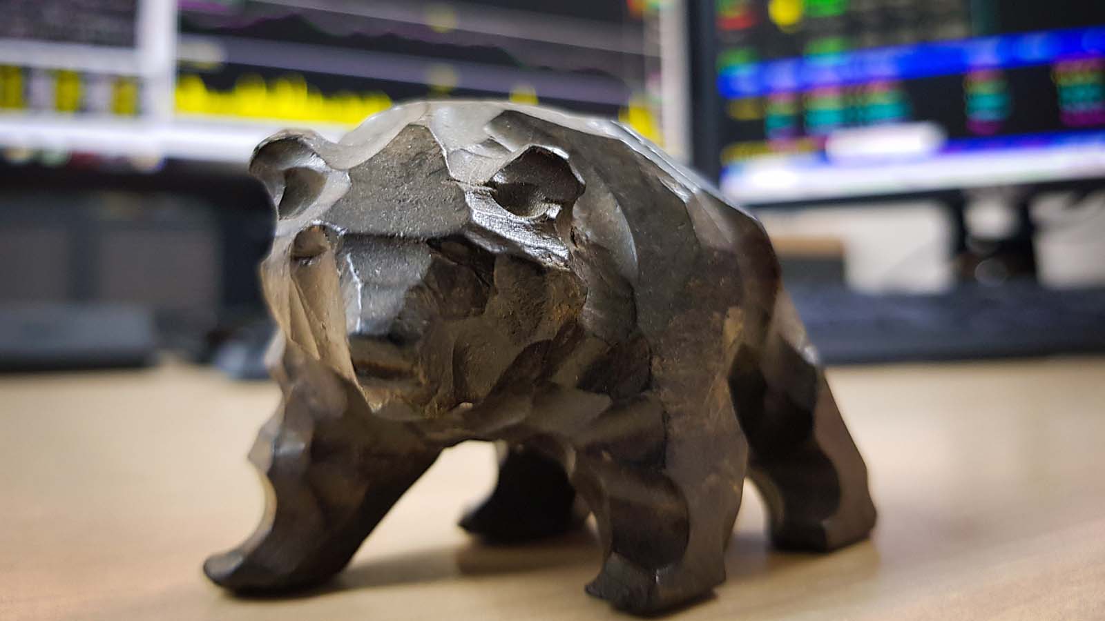 An image of a bear figurine in front of blurred computer screens displaying stock charts