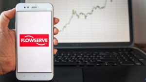 An image of a hand holding a phone displaying the Flowserve Corporation (FLS) logo in front of a laptop displaying a stock chart