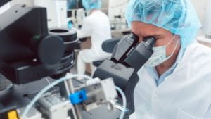 A scientist in medical gear peers through a microscope (IBIO stock)