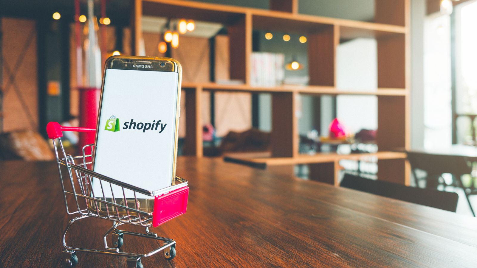Image of a shopping cart toy on a wooden desk carrying a mobile phone that features the Shopy (SHOP) logo on it