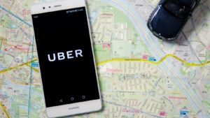 The Uber (UBER) logo is displayed on a smartphone on top of a map background representing UBER Stock.
