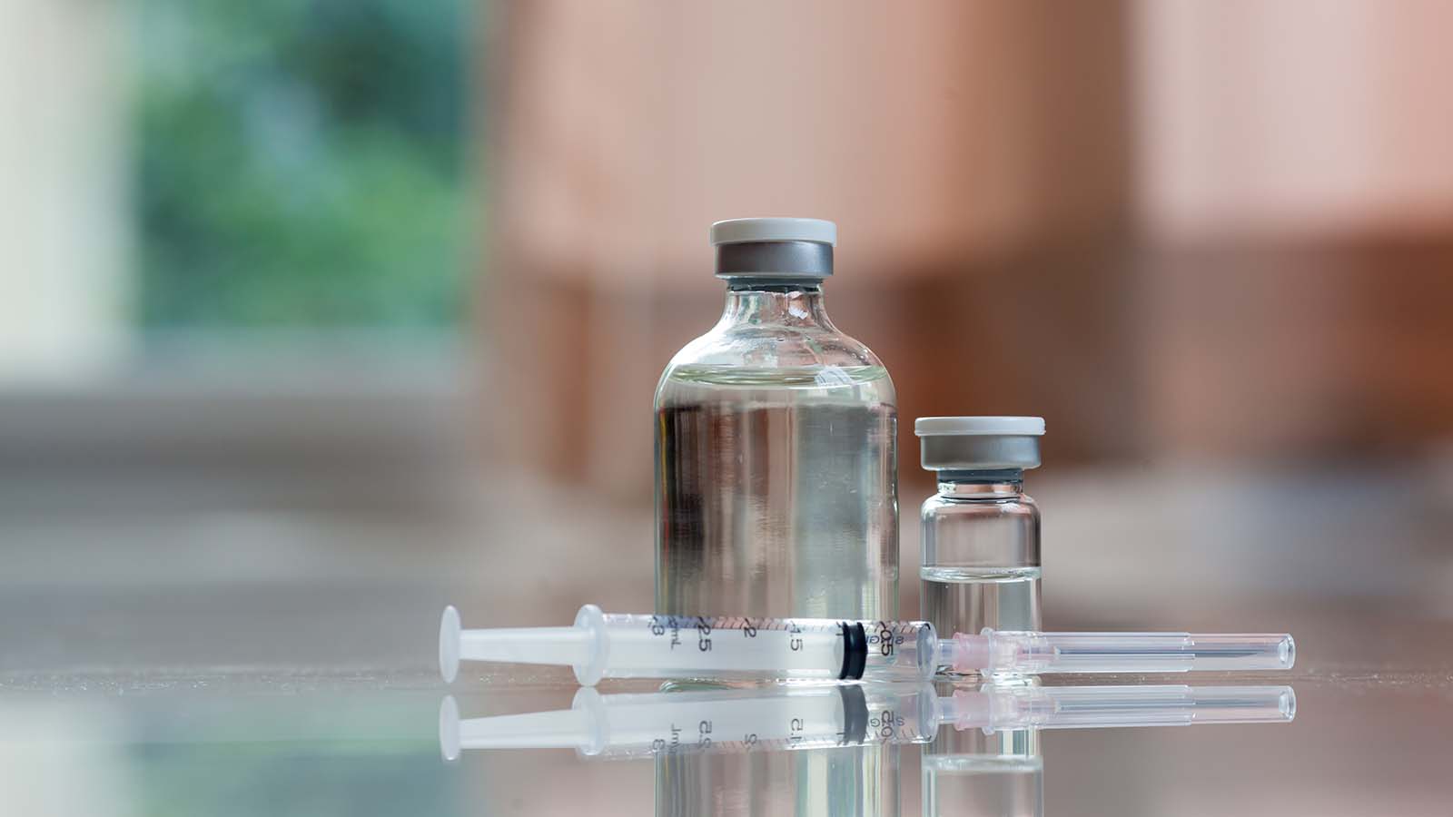 A needle rests on a reflective table next two clear glass vials of a clear liquid.