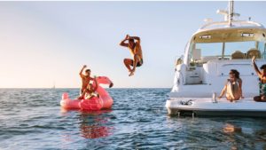 A person jumping off a boat into the water next to their friend who is floating on a flamingo-shaped float