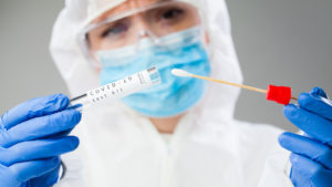 stocks to sell A Medical healthcare technologist holding COVID-19 swab collection kit, wearing white PPE protective suit mask gloves, test tube for taking OP NP patient specimen sample,PCR DNA testing protocol process