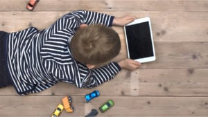 a kid laying on a floor playing with a tablet instead of toy cars that sit next to him