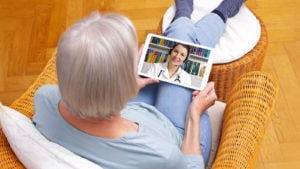 A woman in a wicker chair looking at a doctor on a tablet, chatting.