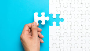 A hand putting the last piece of a white puzzle in place against a blue backdrop.