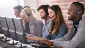 Five young customer support specialists sit in a row at computers with headsets on.