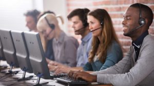 SPRT stock: Five young customer support specialists sit in a row at computers with headsets on.