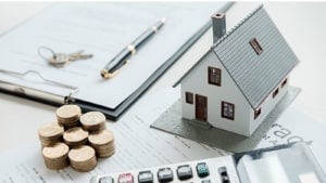 miniature home next to pen, pad of paper, calculator and coins on a desk